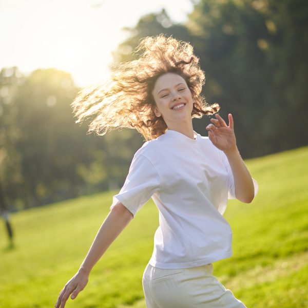 A person with curly hair, wearing a white outfit, is smiling and running through the green park on a sunny day, radiating vitality.