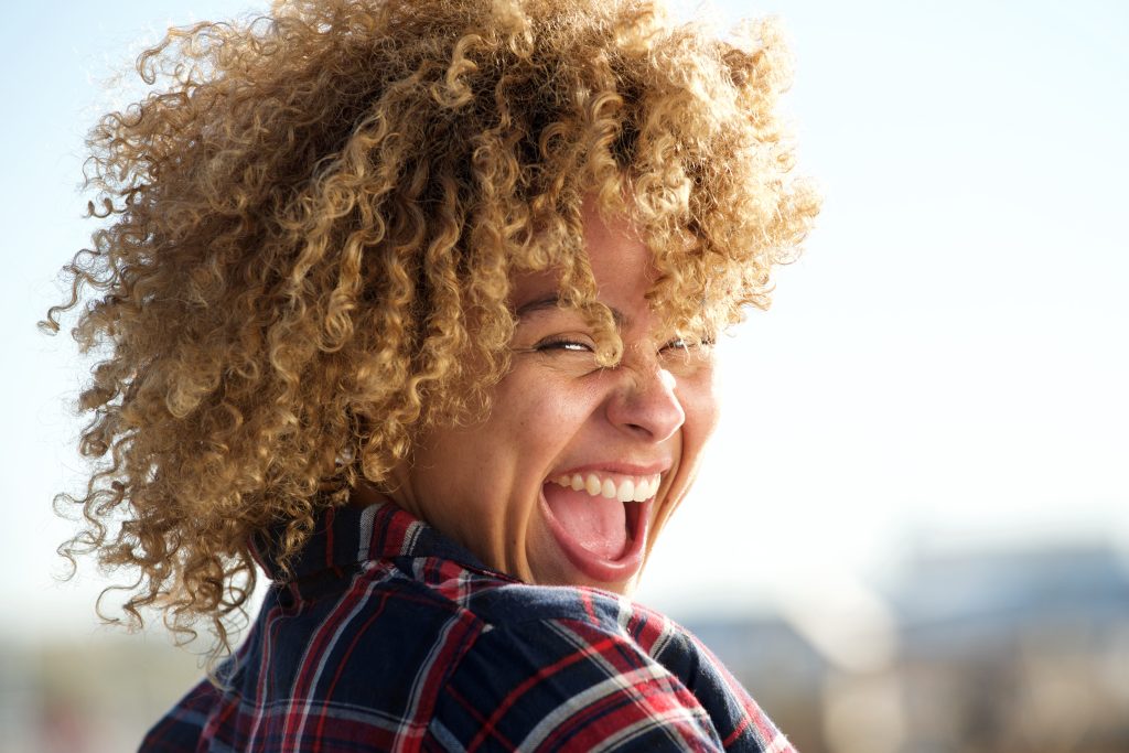 A person with curly hair exudes vitality, smiling widely while wearing a plaid shirt under the bright sunlight.
