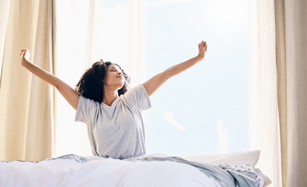 A person exudes vitality as they stretch with arms raised while sitting on a bed in a sunlit room.