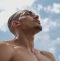 A shirtless man looks upwards towards the sky, with clouds and blue sky in the background.