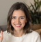 Woman with shoulder-length brown hair smiling, wearing a white sweater. Indoor setting with a green plant in the background.