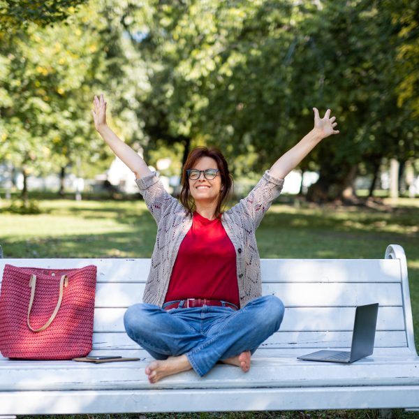 A woman sitting cross-legged on a white bench in a park, smiling with arms raised, enjoys a moment of calm by practicing basic breathing techniques. Beside her sit a red bag and an open laptop, hinting at a day intertwined with both serenity and modern stress.