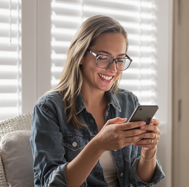 A woman wearing glasses and a denim jacket is smiling while looking at a smartphone, with window blinds in the background.