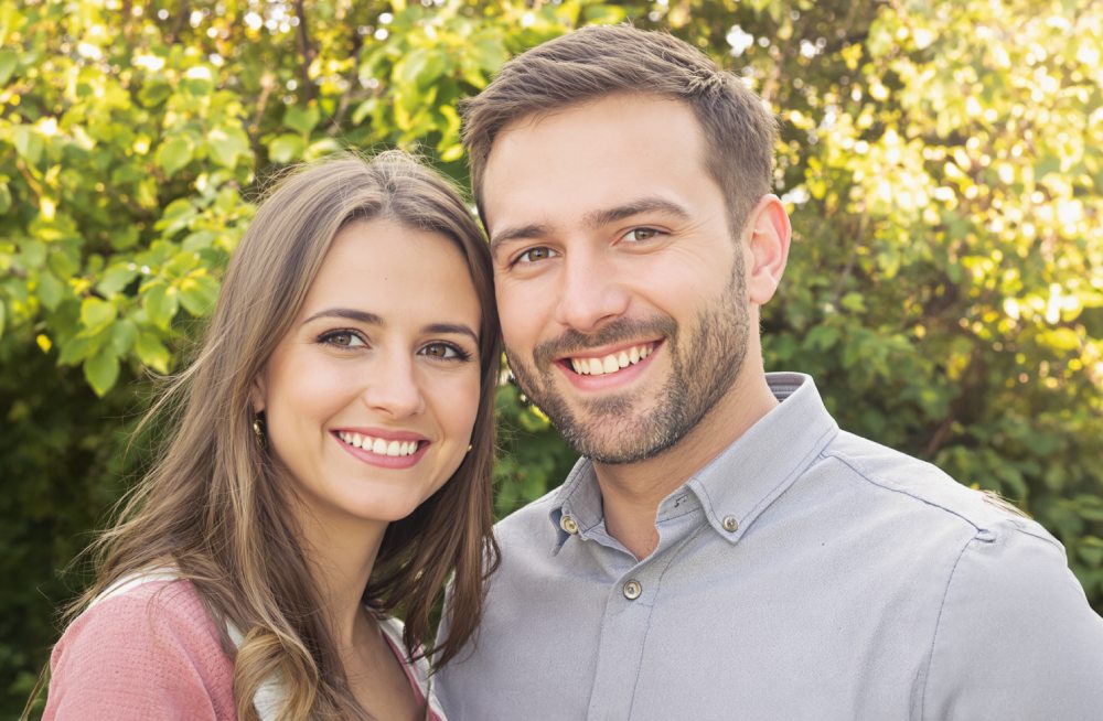 A couple is smiling at the camera outdoors, with green foliage in the background.