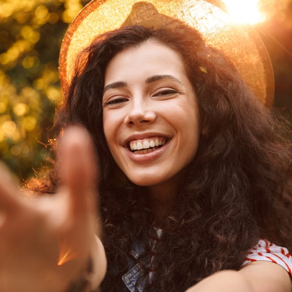 A person with long curly hair smiles warmly, reaching towards the camera. Sunlight filters through a hat, enhancing the sense of wellbeing in this serene outdoor setting—like a professional guide leading you to embrace health and happiness.