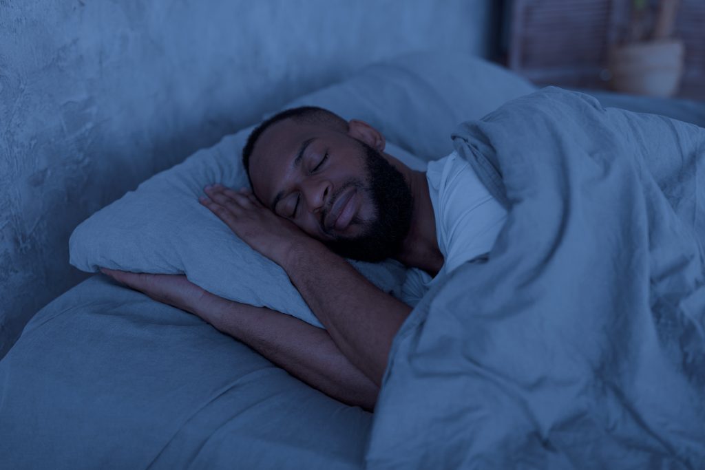 A person with a beard sleeps peacefully, resting their head on a pillow and covered with a blanket in a dimly lit room, embracing the essence of wellbeing.