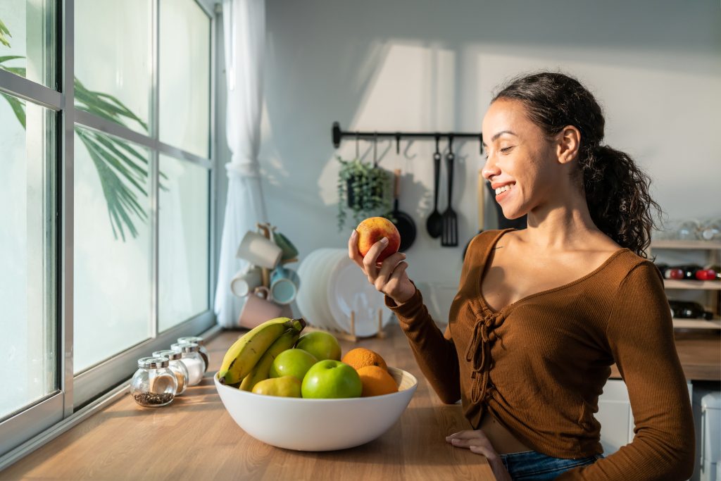 A person stands in a kitchen, smiling at a peach they hold, symbolizing better health. A bowl of assorted fruits rests on the wooden counter. Sunlight streams through a window with plants outside, offering an idyllic backdrop to optimise your daily wellbeing.